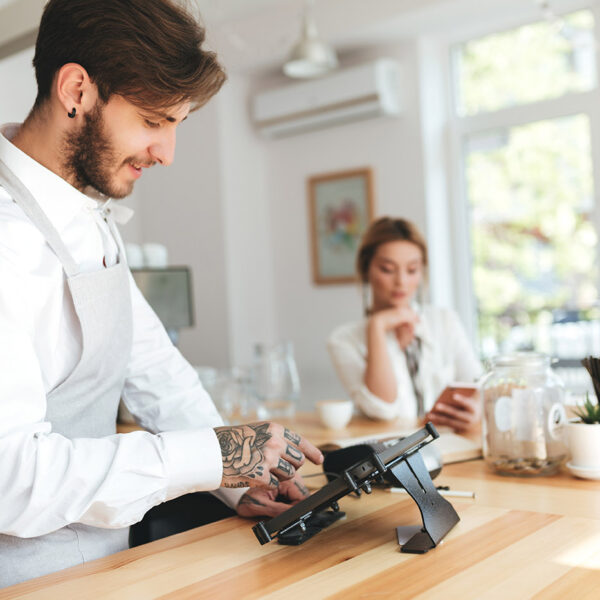 Smiling barista in apron using cash counter while girl at the counter using smartphone